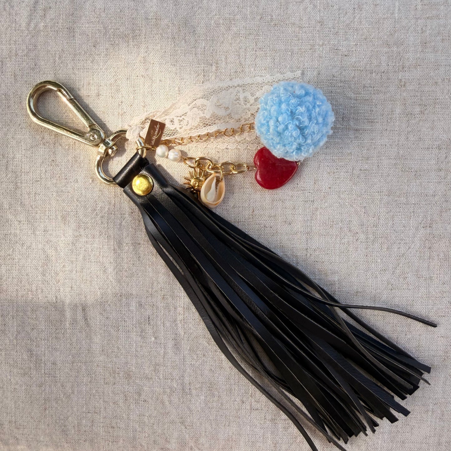 A black leather tassel keychain with a gold-tone lobster clasp, featuring a light blue pom-pom, a red heart charm, and small gold and pearl accents, displayed on a textured beige linen background.